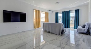 A spacious room with a flat-screen TV mounted on the wall, a bed with a grey blanket, a small sofa with a grey cushion, and a window with blue curtains. - Prime Swieqi 6BR w/ Private Pool & Sundeck (Swieqi)