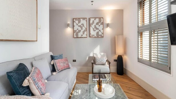 A modern living room with a sofa, armchair, and coffee table, featuring artwork and a lamp. - Modern Fulham 1-bedroom Apartment (London)