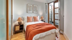 A bedroom with a bed, two bedside lamps, a nightstand, a window with shutters, and two framed pictures on the wall. - Modern Fulham 1-bedroom Apartment (London)