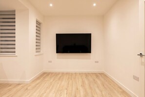 Ein Zimmer mit flachbildfernseher an der Wand, hellem Holzboden und einem Fenster mit Jalousien.