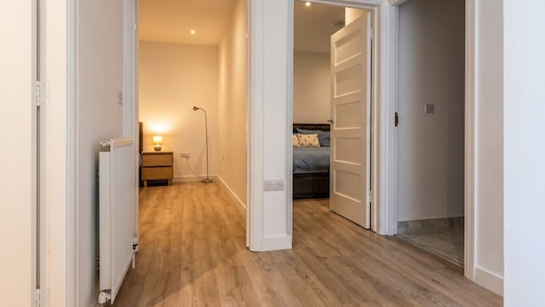 A hallway with wooden flooring, a radiator, a bedside table with a lamp, and a bed with a striped bedspread. - Newly refurbished London base with Jubilee line on your doorstep (Greater London)