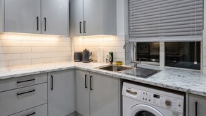 A modern kitchen with white cabinets, a Bosch washing machine, and a window with blinds. - Newly refurbished London base with Jubilee line on your doorstep (Greater London)