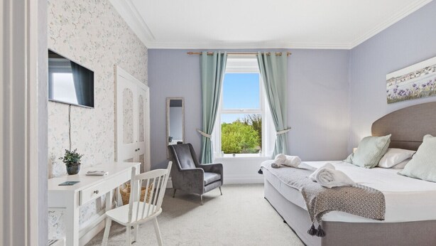 A bedroom with a bed, a desk, a chair, a window with curtains, and a view of greenery outside.