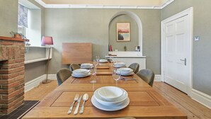 A dining room with a wooden table set for four, a brick fireplace, and a view of the kitchen through an archway.