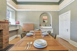 A dining room with a wooden table set for four, a brick fireplace, and a view of the kitchen through an archway.