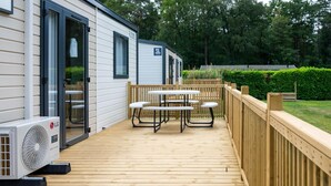 A mobile home with a deck, outdoor table, and chairs, surrounded by a wooden fence and greenery.