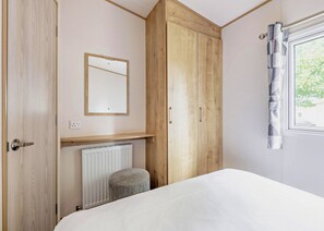 A bedroom with a bed, a wooden wardrobe, a radiator, a small stool, a mirror, and a window with curtains. - Gold 3 Bedroom Caravan, Sleeps 8. With decking (Clacton-on-Sea)
