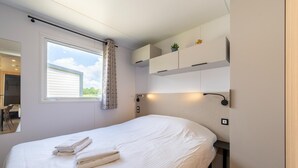 A neatly made bed with towels, a window with curtains, built-in cabinets, and a wall-mounted lamp.