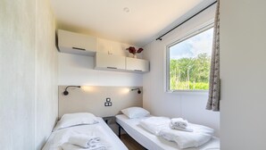 A room with two single beds, a small shelf with decorative items, a window with curtains, and a wall-mounted light fixture. - Comfortable caravan in a green setting (Lembeke (Kaprijke))
