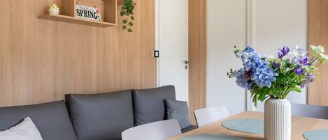 Ett modernt rum med en soffa, ett matbord med underlägg, en vas med blommor och en hylla med en skylt och en växt.