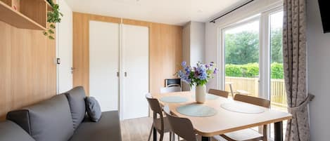 A modern room with a sofa, dining table, chairs, a vase with flowers, and a view of greenery through large windows.