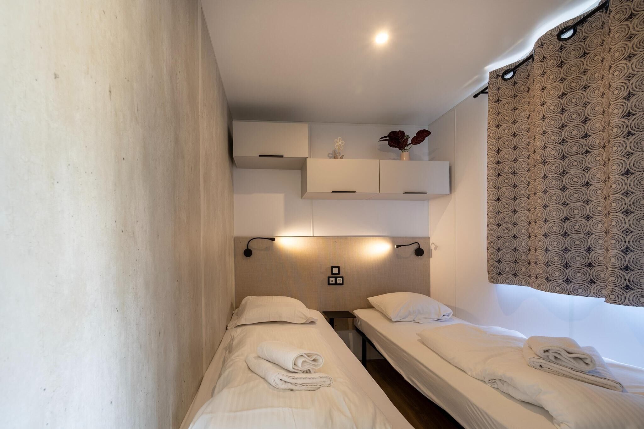 Quarto de hotel com duas camas de solteiro, uma pequena mesa de cabeceira, uma prateleira fixada na parede com objetos decorativos e cortina estampada.