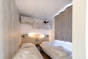 A hotel room with two single beds, a small bedside table, a wall-mounted shelf with decorative items, and a window with patterned curtains.