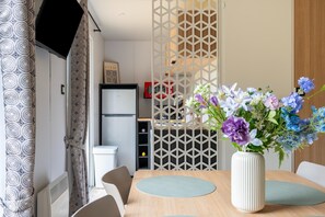A dining area with a table, chairs, a vase with flowers, a refrigerator, and a patterned curtain.