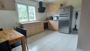 A kitchen with a wooden table, chairs, a refrigerator, a microwave, and a window with a view of trees. - Gîte in the heart of nature, peace and quiet guaranteed, animals welcome. (Diges)