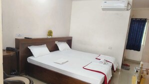 A single-bed room with a wooden headboard, white bedding, a small desk with a chair, an air conditioning unit, and a window with a curtain. - Binayak Palace OD (Kendrapara)