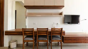 A modern kitchen with a wooden dining table, three chairs, a TV mounted on the wall, and a built-in cabinet.