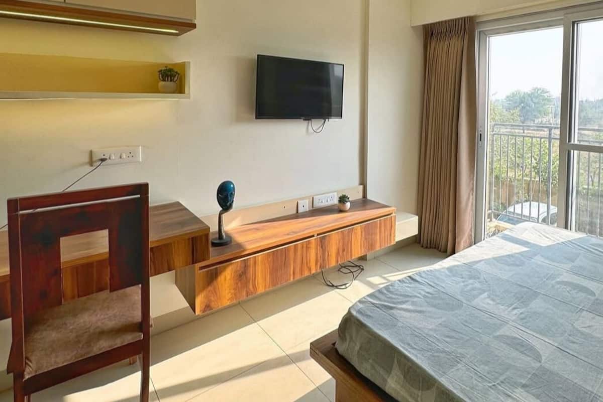 Un dormitorio con cama, escritorio, silla, televisor y balcón con cortinas.