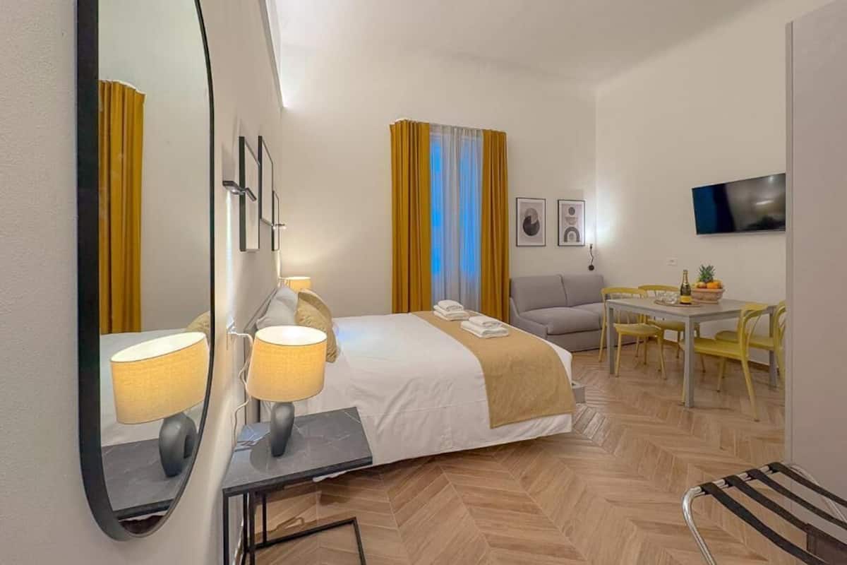 A hotel room with a bed, bedside lamps, a desk, a chair, a table, a television, and curtains.