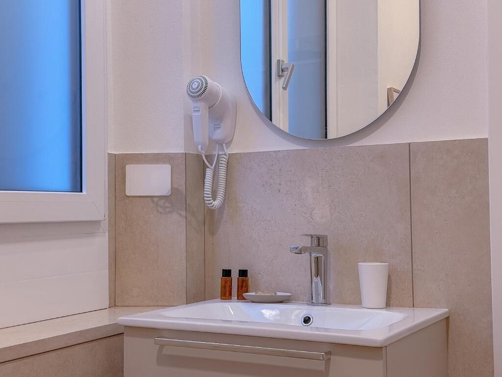 A bathroom with a white sink, a round mirror, a hairdryer, and a window.