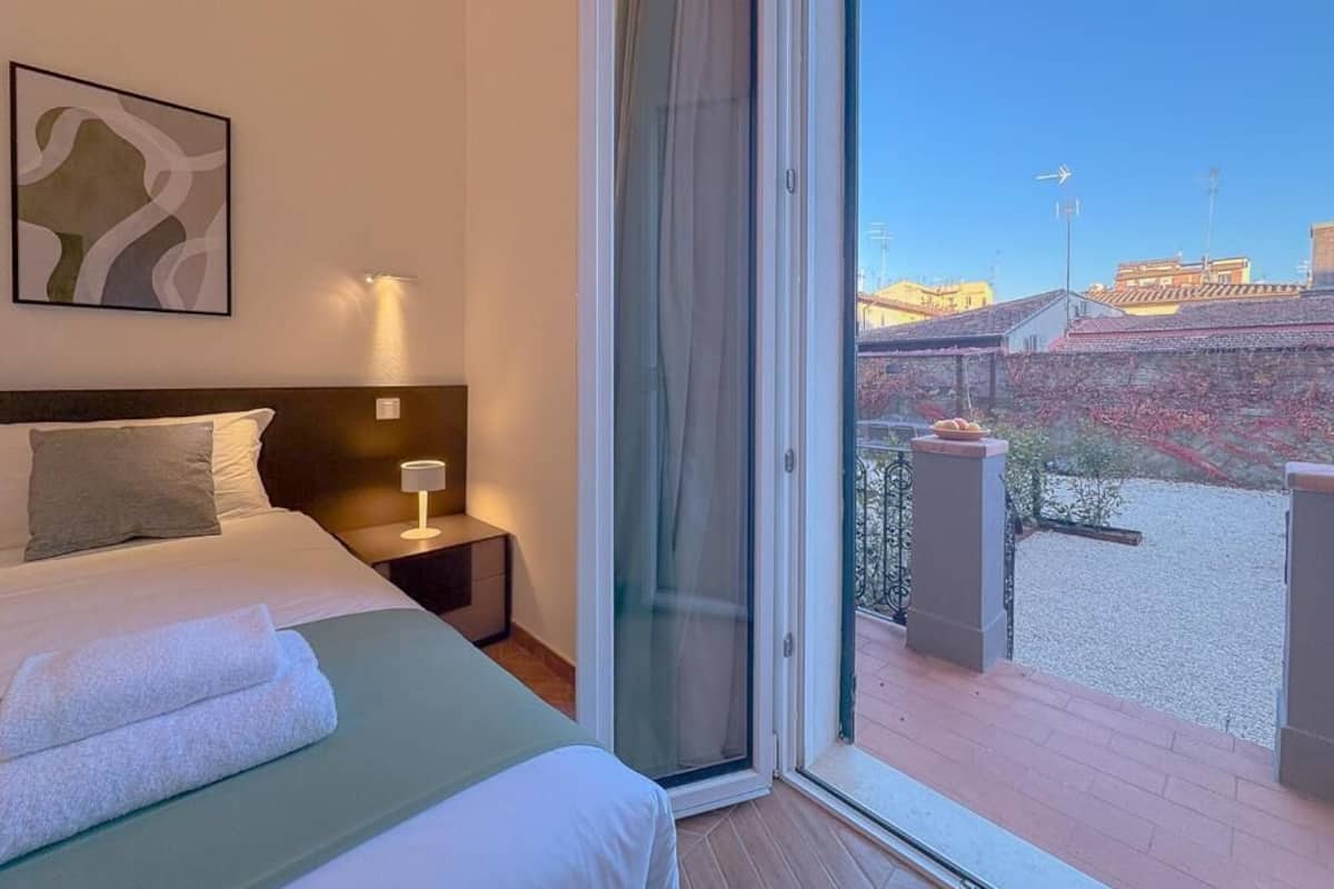 A hotel room with a bed, a nightstand, a lamp, and a view of a cityscape through a glass door.