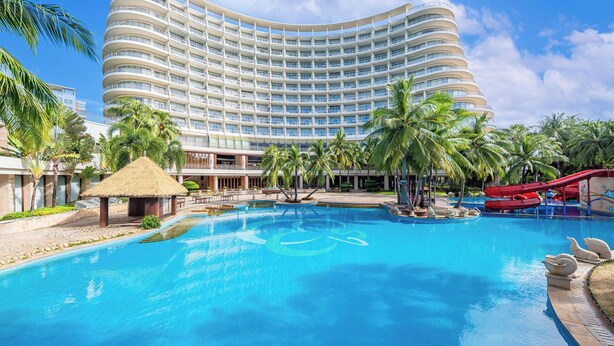 Un hotel grande con fachada curva, múltiples balcones, piscina central, palmeras y un tobogán rojo.