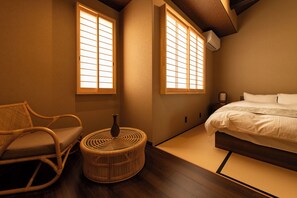 Un dormitorio de estilo japonés tradicional con suelo de tatami, una cama baja, una silla de madera y una pequeña mesa redonda.
