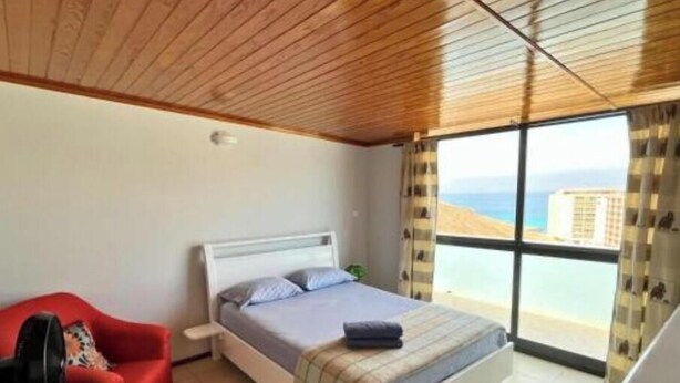 A bedroom with a bed, a red chair, a standing fan, and a large window with a view of the sea. - Dorade Double Room – Cozy Stay at Alto do Fortim B&B (Mindelo)