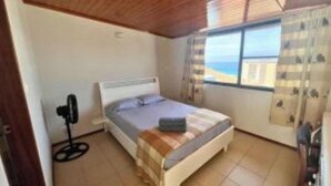 A bedroom with a bed, a window, a fan, and a view of the beach. - Dorade Double Room – Cozy Stay at Alto do Fortim B&B (Mindelo)
