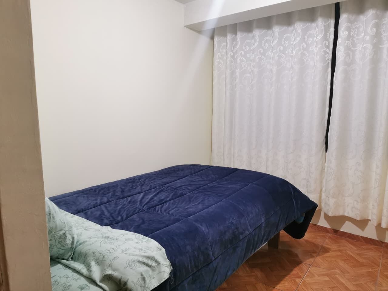 A small, single-bed room with a blue bedspread, a white curtain, and a wooden floor.