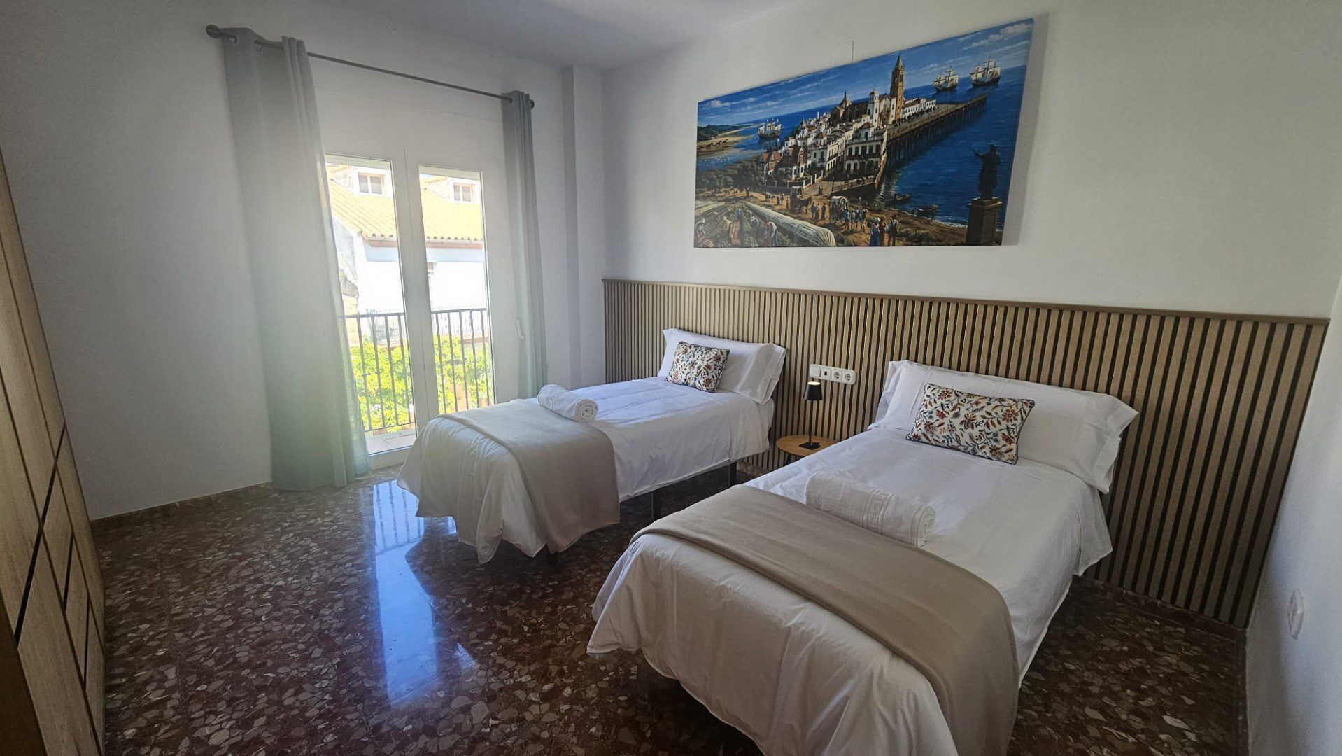 Habitación de hotel con dos camas, un ventanal con cortinas, un cuadro de un pueblo costero y suelo de baldosas.