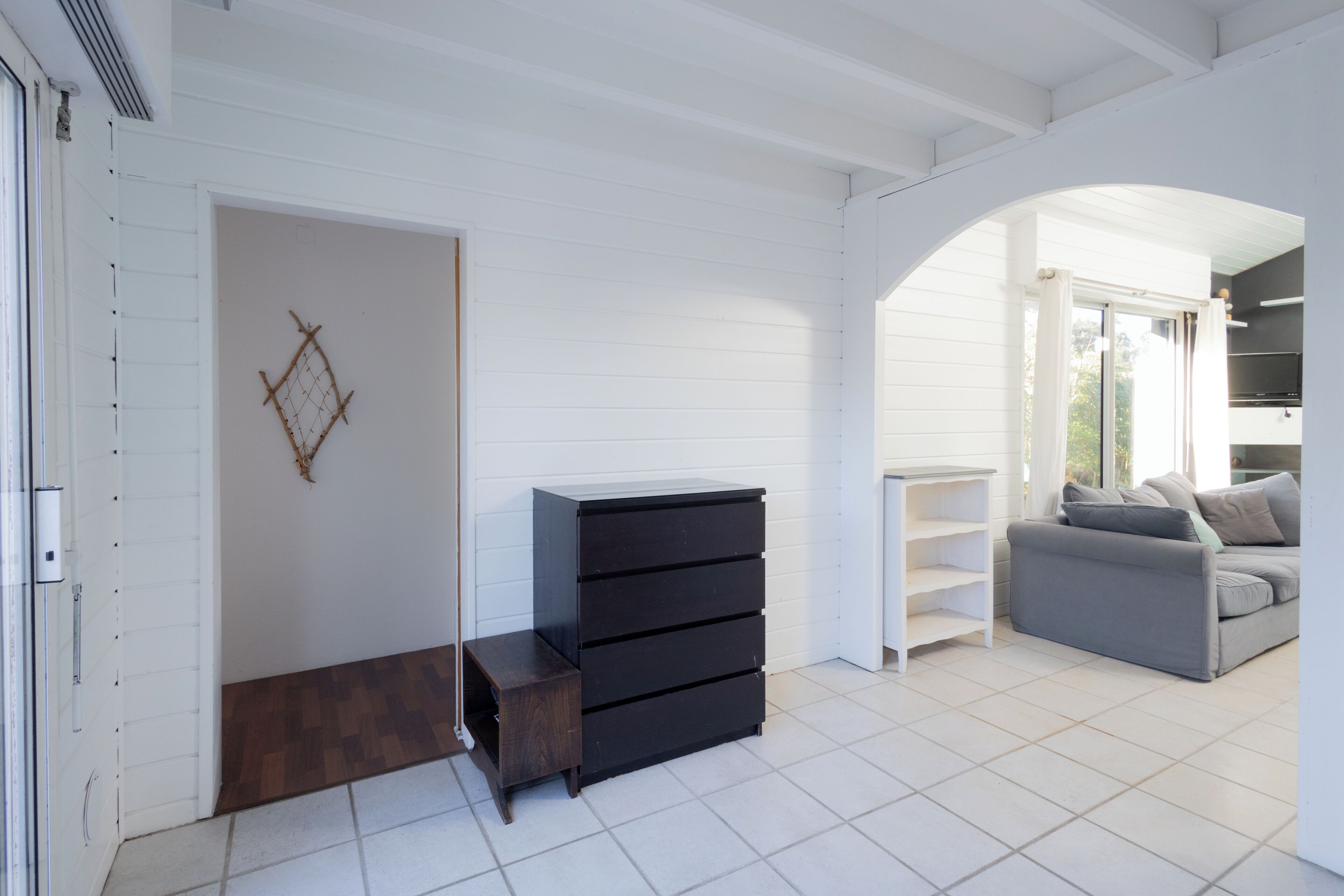 Una habitación con pared blanca, un banco de madera, un aparador negro, un sofá gris y un estante blanco.