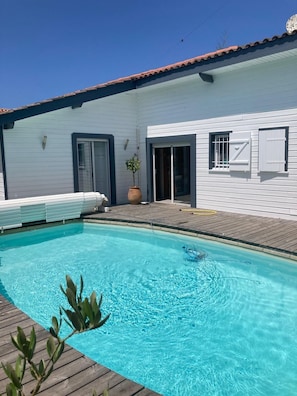 Une piscine avec une terrasse en bois, un bâtiment blanc doté d’un toit en tuiles et une plante en pot.