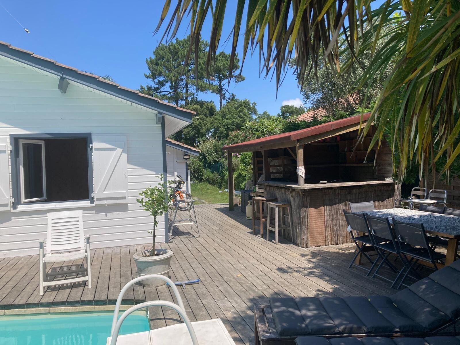 Une piscine avec une terrasse en bois, un espace barbecue et une maison blanche avec des volets.