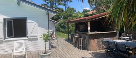Une piscine avec une terrasse en bois, un espace barbecue et une maison blanche avec des volets.