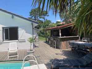 Une piscine avec une terrasse en bois, un espace barbecue et une maison blanche avec des volets.