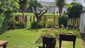 Una terraza de madera con plantas en macetas, un banco y un césped con una valla al fondo.