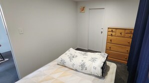A small bedroom with a single bed, a wooden dresser, and a door. - The Nest - Dive in to Lloyd Elsmore Park for infinite opportunities (Manukau City)