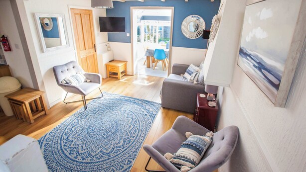 Een woonkamer met een blauw kamerkleed, een grijze bank, een houten bijzettafeltje, een witte fauteuil en een aan de muur bevestigde televisie.