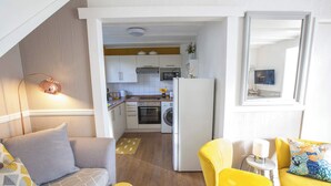 Un espace de vie compact comprenant un coin cuisine, un canapé, un fauteuil jaune et une petite table avec une lampe.