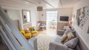 Un salon avec un canapé, un fauteuil jaune et un tapis à motifs. Un téléviseur est fixé au mur, et un tableau est accroché au mur.