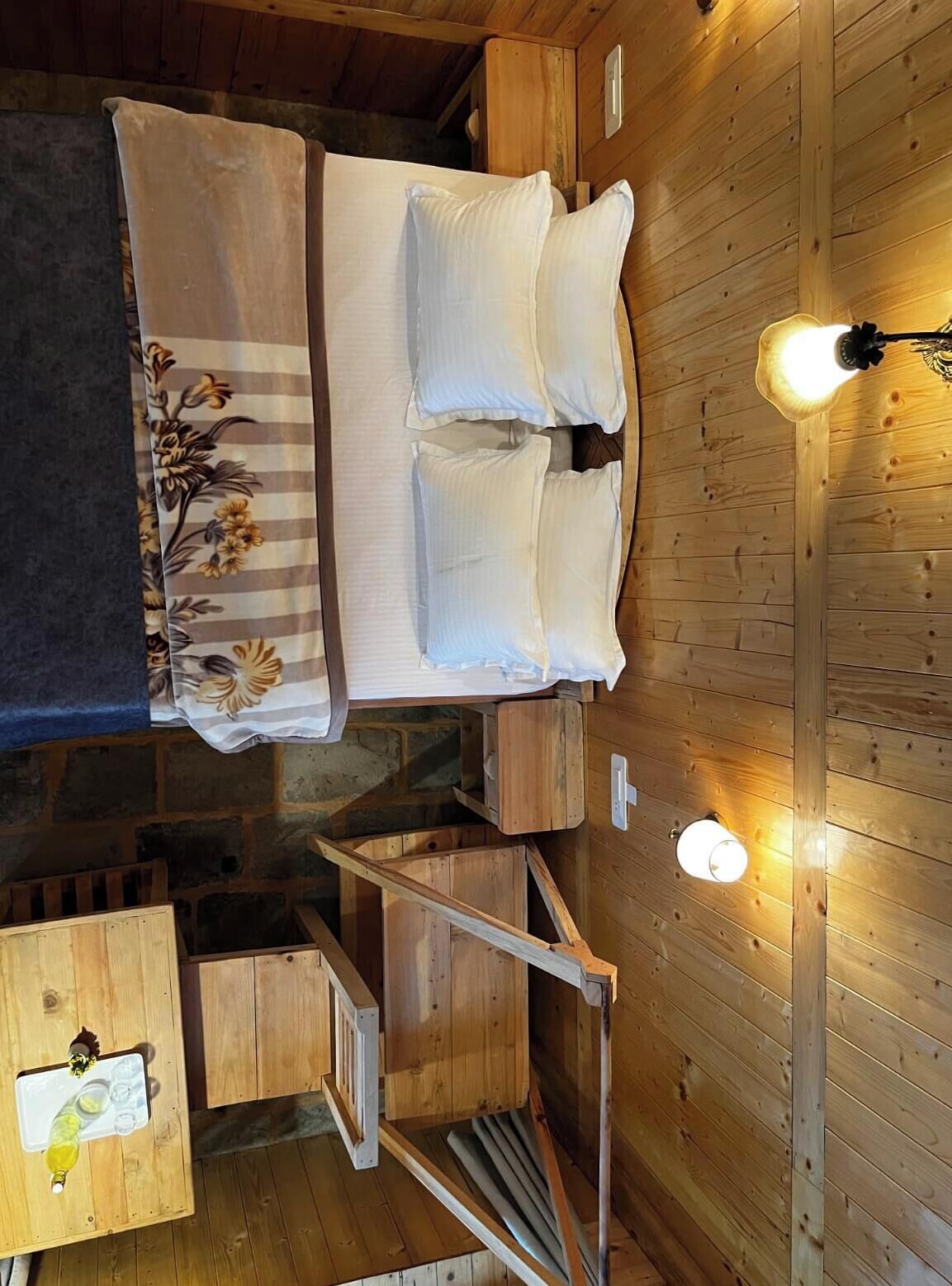 A wooden cabin interior with a bed, two bedside lamps, a small table with a bottle, and a chair.