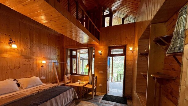 Een houten hutje met een bed, een tafel en een stoel. Er is een balkon met uitzicht op de natuur.