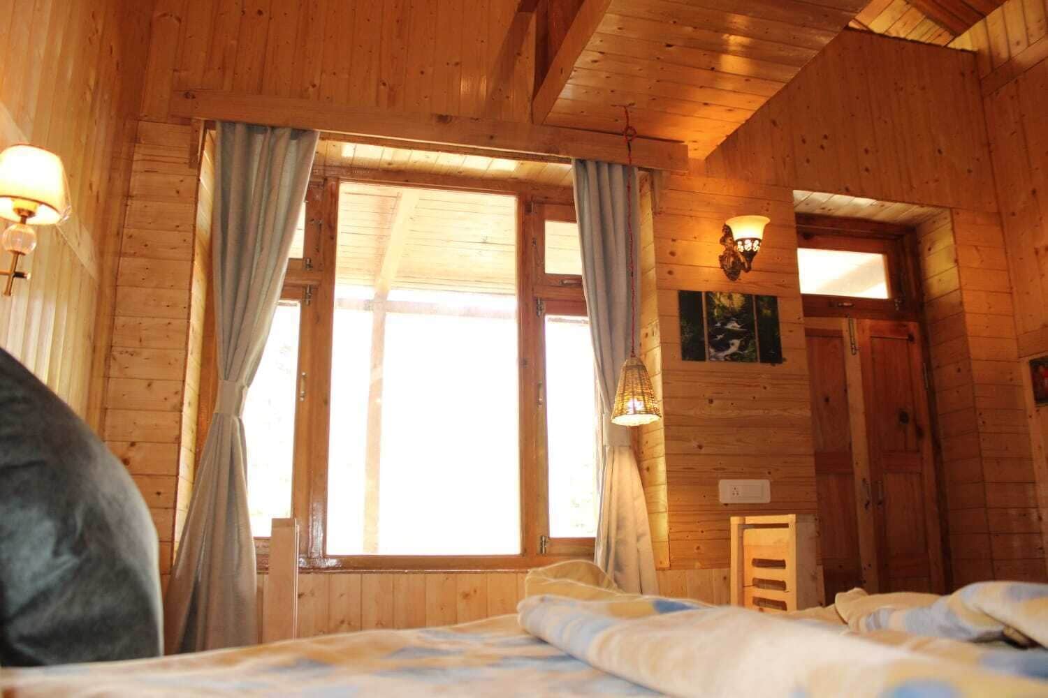 A wooden cabin room with a bed, curtains, a window, and a wall-mounted light fixture.