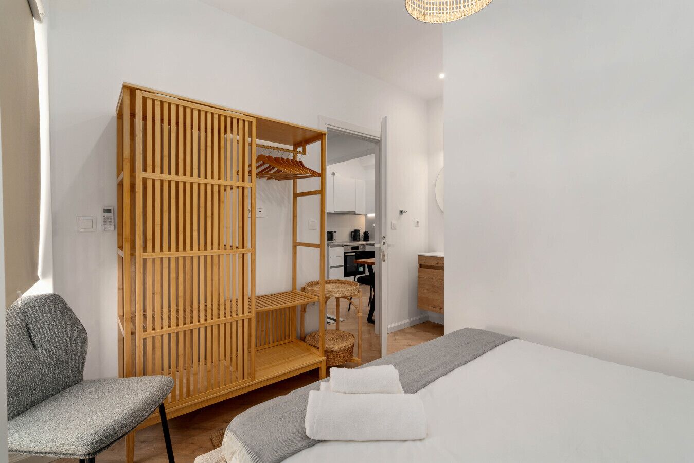 A bedroom with a bed, a chair, a wooden wardrobe, and a kitchenette in the background.