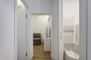 Un pasillo estrecho con suelo de madera, una puerta blanca a la izquierda, un mueble blanco con lavabo y toallero a la derecha, y un sofá marrón al fondo.
