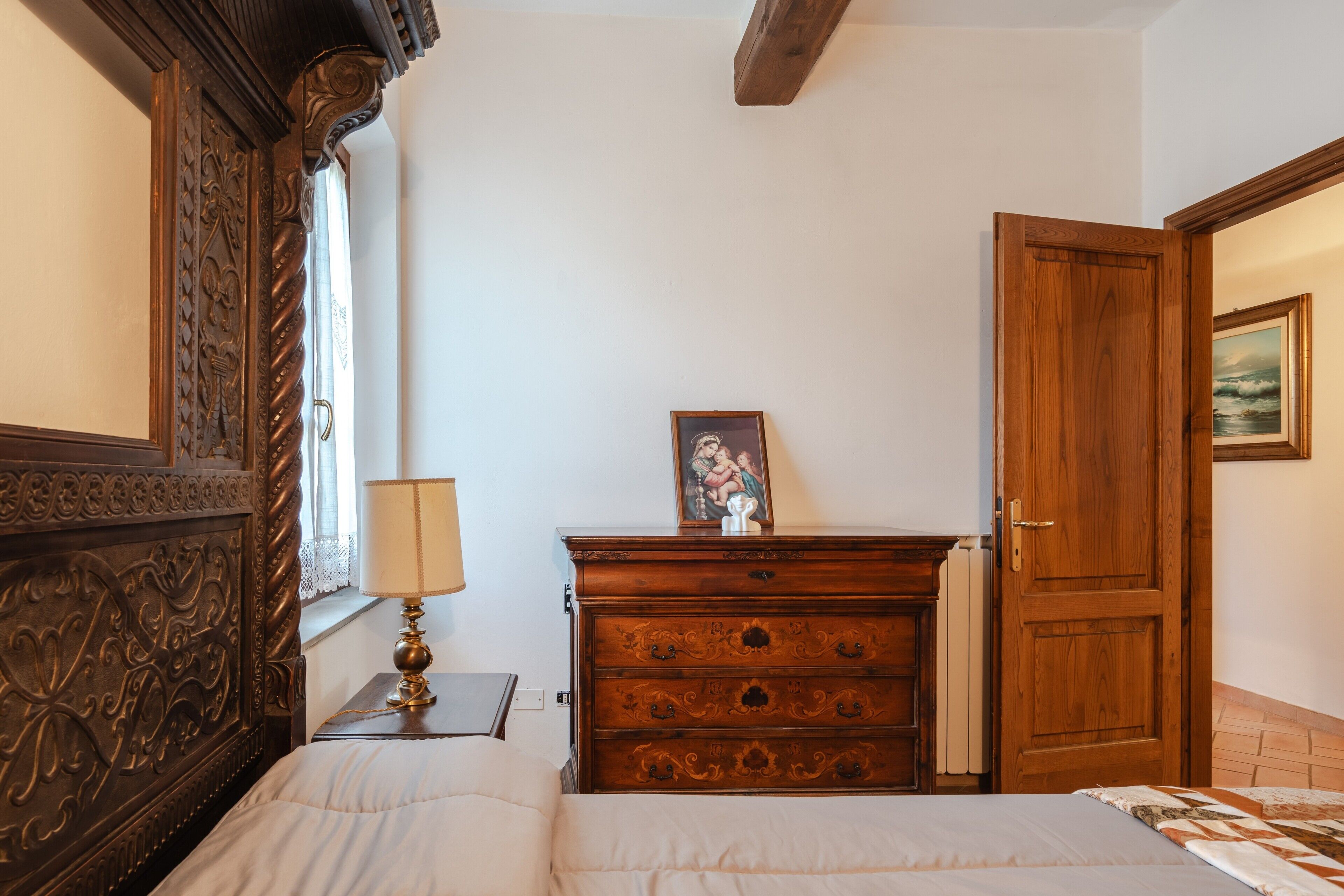 Ein Schlafzimmer mit einem Bett, einer hölzernen Kommode, einer Lampe, einer Tür und einem Bild an der Wand.