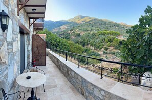 A balcony with a stone wall, a table, and chairs, overlooking a mountainous landscape with trees and a house.
