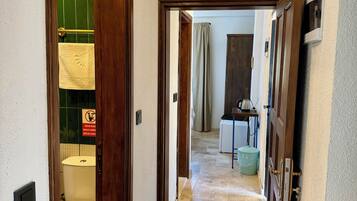 A hallway with a toilet, a trash bin, and a door leading to another room.
