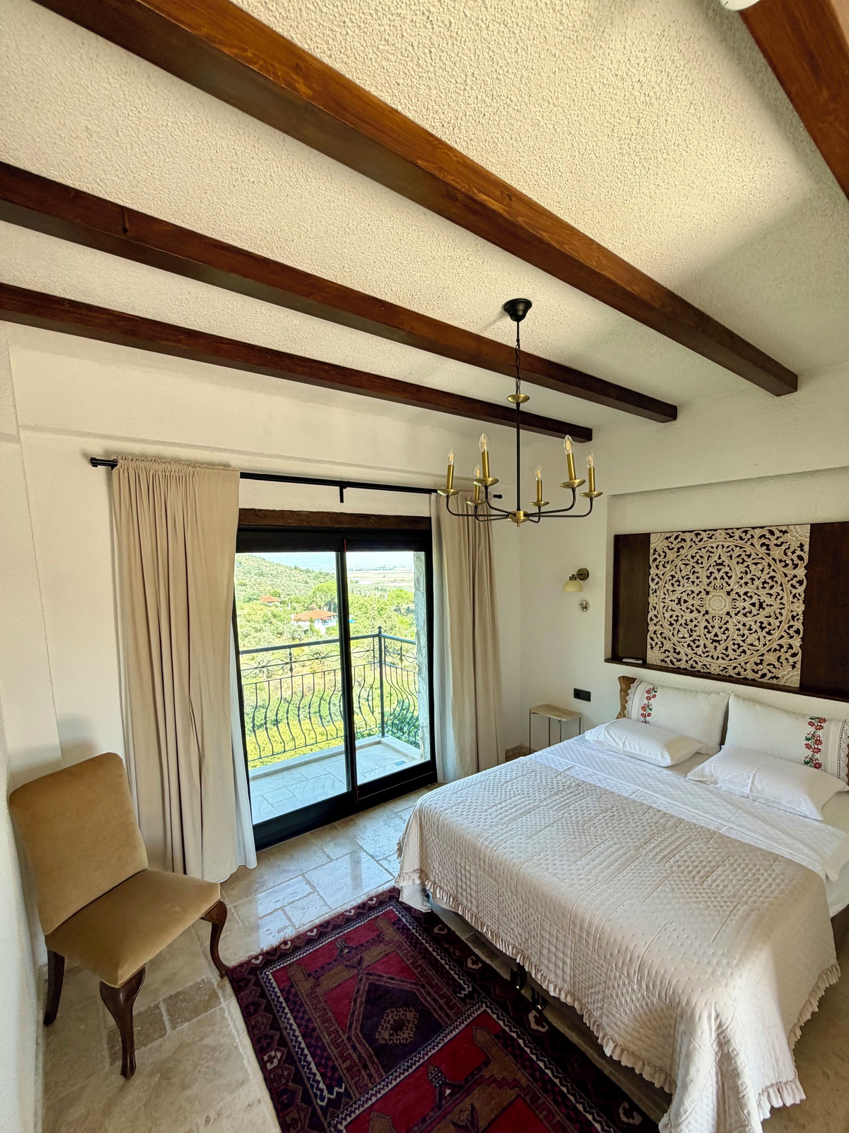 Un dormitorio con una cama grande, una silla, un tapete estampado, una lámpara de araña y vistas a zonas verdes por la ventana.